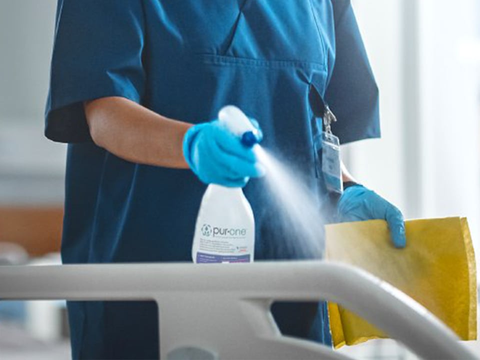 Gloved hand sprays medical bed with bottle of PurOne disinfectant 